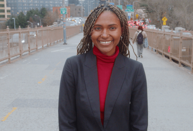 A young woman in business clothing stands on an NYC bridge and smiles at the camera